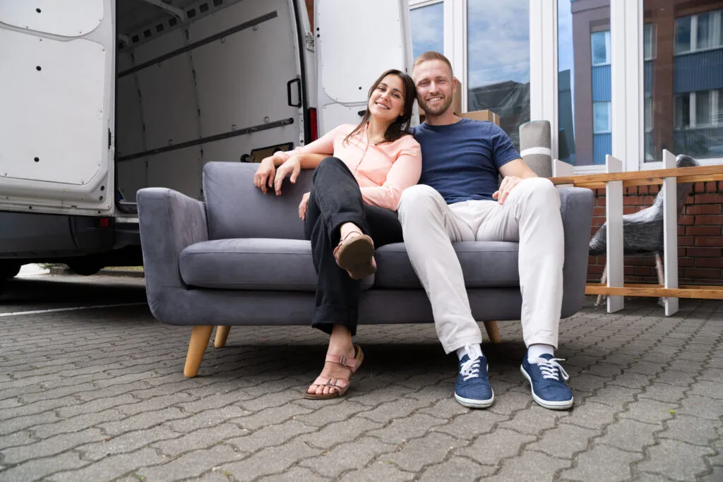 Paar sitzt nach dem Möbeltransport in Wien auf einem grauen Sofa vor dem offenen Transporter