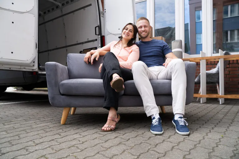 Paar sitzt nach dem Möbeltransport in Wien auf einem grauen Sofa vor dem offenen Transporter